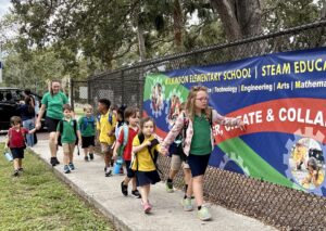 Elementary school students walk along a sidewalk near a Wilkinson Elementary School banner under adult supervision.
