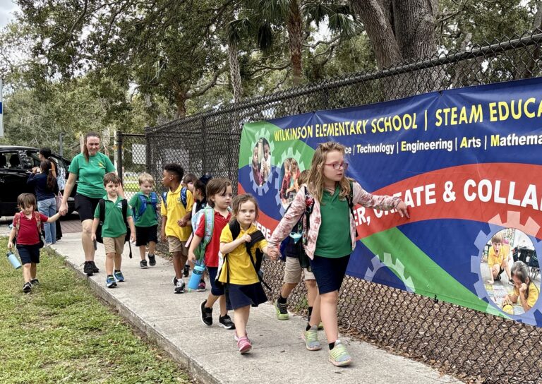 Elementary school students walk along a sidewalk near a Wilkinson Elementary School banner under adult supervision.
