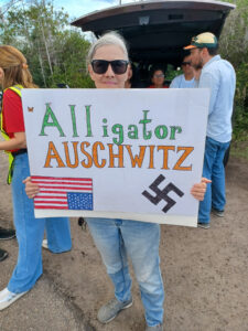 A demonstrator holds a sign that reads "Alligator Auschwitz" with an upside-down US flag and a swastika.