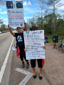 Roxana Torres and another demonstrator holding signs. Torres' sign reads, "My husband been detained since October 30. The children need their father". The other demonstrator's sign reads "One single toilet roll for 30 person. There are a lot of people inside that have ACOL and other various illnesses. They are not given access to their medications; not insulin for diabetics".