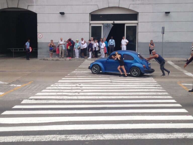 Two people push a broken-down car down a city street while several others spectate.
