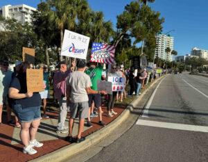 Protesters stand along a downtown sidewalk holding anti-ICE signs such as "I like my ice crushed!" and "Imagine fearing diversity but not dictatorship".