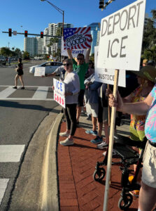 Protesters stand along a downtown sidewalk holding anti-ICE signs such as "Deport ICE," "Remember Good!" and "ICE murdered Good".