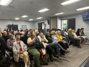 A full crowd sits in a board meeting room, many members with raised hands.