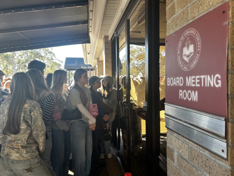 A crowd gathers at the entrance of a building with a sign that reads "Board Meeting Room" with a Sarasota County School Board seal.