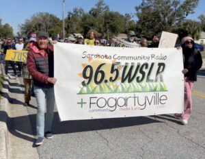 Demonstrators hold a banner that reads: Sarasota Community Radio 96.5 LPFM WSLR + Fogartyville Community, Media & Arts Center