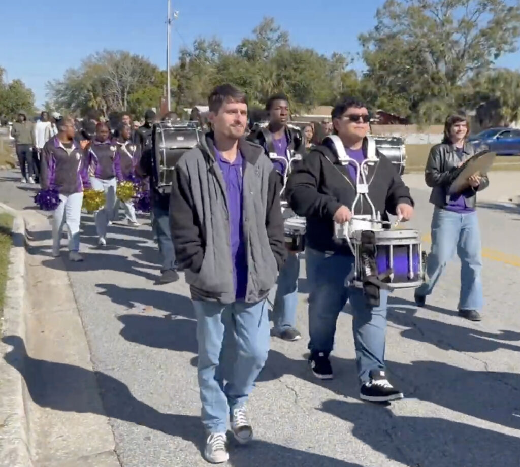 Marching band percussionists with purple and black uniforms proceed down the street.