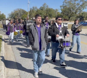 Marching band percussionists with purple and black uniforms proceed down the street.