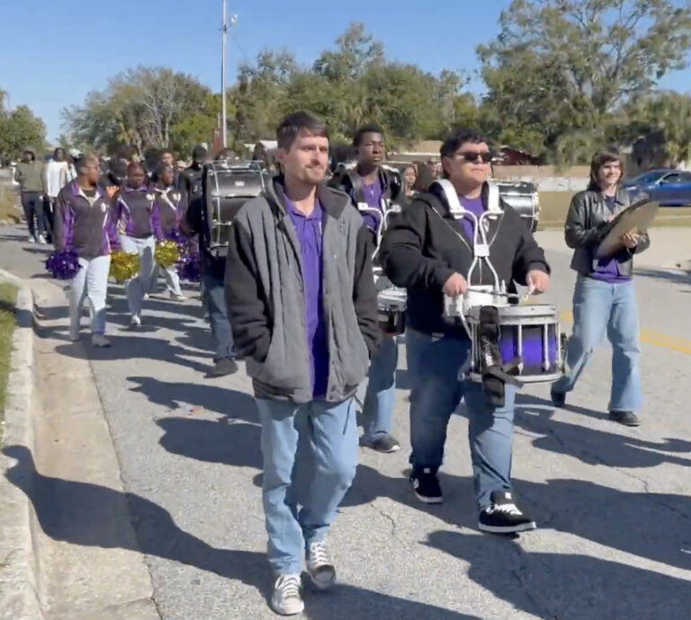 Marching band percussionists with purple and black uniforms proceed down the street.