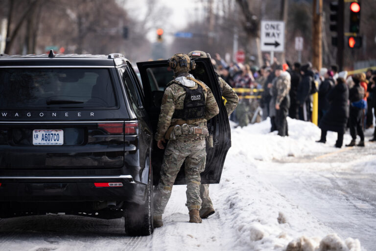 An uniformed police officer lingers by the open door of a black SUV in the snow amid a crowd of demonstrators.