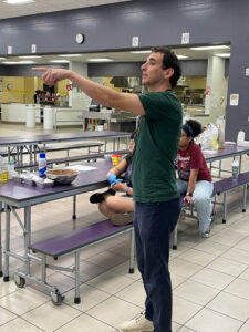 Andrew Grossman points and addresses someone off-camera in a school cafeteria.