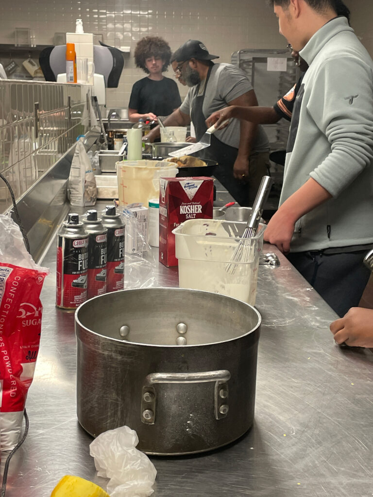 A pot sits on the counter in a busy kitchen where students work alongside Chef G.