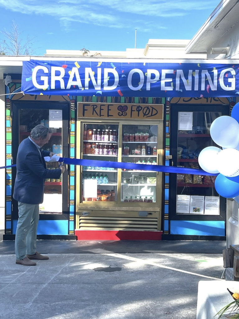 A person cuts a ribbon in front of a community fridge with "Free Food" painted on it and a colorful "GRAND OPENING" banner.