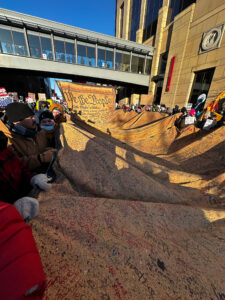 Demonstrators on a crowded city street work together to hold a very long banner with innumerable signatures, one end of which appears to have the beginning of the Constitution written on it.