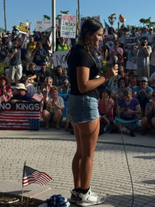 A speaker addressing a crowd of demonstrators holding signs with messages as "No kings; impeach, remove, convict".
