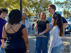 Demonstrators talk amongst themselves. One holds a sign that reads "Keep school board meetings @ 6 PM!"