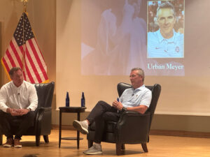 Urban Meyer reclining in a chair on stage in front of a presentation slide with his portrait and name.