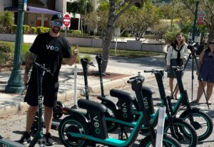 A person in a Veo t-shirt and hat gives a thumbs-up next to several stationary Veo scooters and bikes.