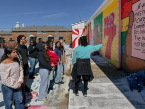 A speaker gestures to a mural with graphic text that reads "Mar 7" while addressing a crowd.