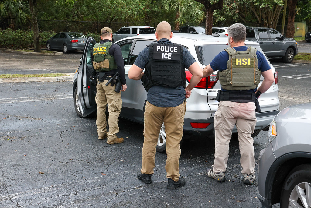 Three ICE officers stand near a vehicle with their backs to the viewer.