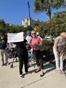 Demonstrators along the sidewalk. One holds a sign that reads "No Kings."