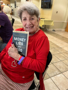 A smiling person seated in a conference hall holds a copy of "Money, Lies, and God" by Katherine Stewart.