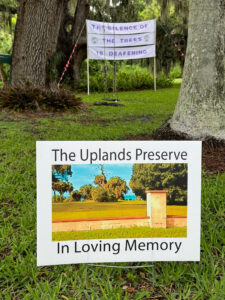 Two signs in a wooded area. The sign in the foreground reads, "The Uplands Preserve / In Loving Memory". The sign in the background reads "The silence of the trees is deafening".