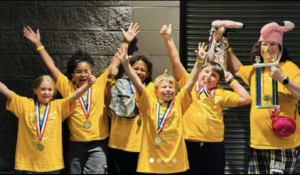 Students in yellow shirts lift their arms in celebration and pose with a first-place trophy.