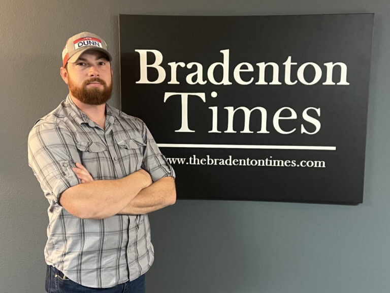 John Dunn posing with arms folded next to a Bradenton Times sign.