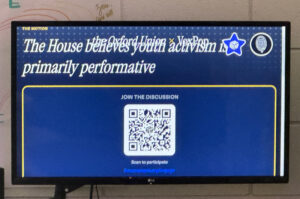 A screen showing a QR code inviting spectators to "join the discussion" and respond to the prompt "The House believes youth activism is primarily performative".
