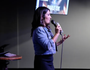Michelle Pozzie speaking into a microphone in front of a screen with text that reads "Charlie Kirk."