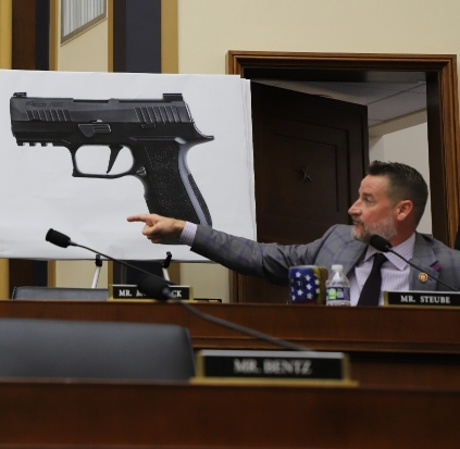 Greg Steube gesturing in front of a big presentation slide of a handgun.