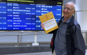 Douglas Dixon smiling and holding an envelope in an airport.