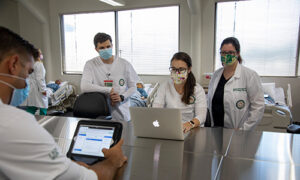 Nursing students wearing face masks crowd around a laptop. Patients lie in hospital beds behind them.
