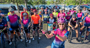 A group of cyclists poses for a photo. The cyclist closest to the camera holds up an award.