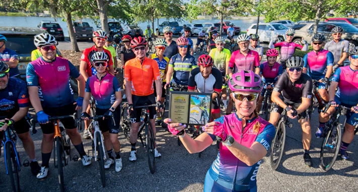 A group of cyclists poses for a photo. The cyclist closest to the camera holds up an award.