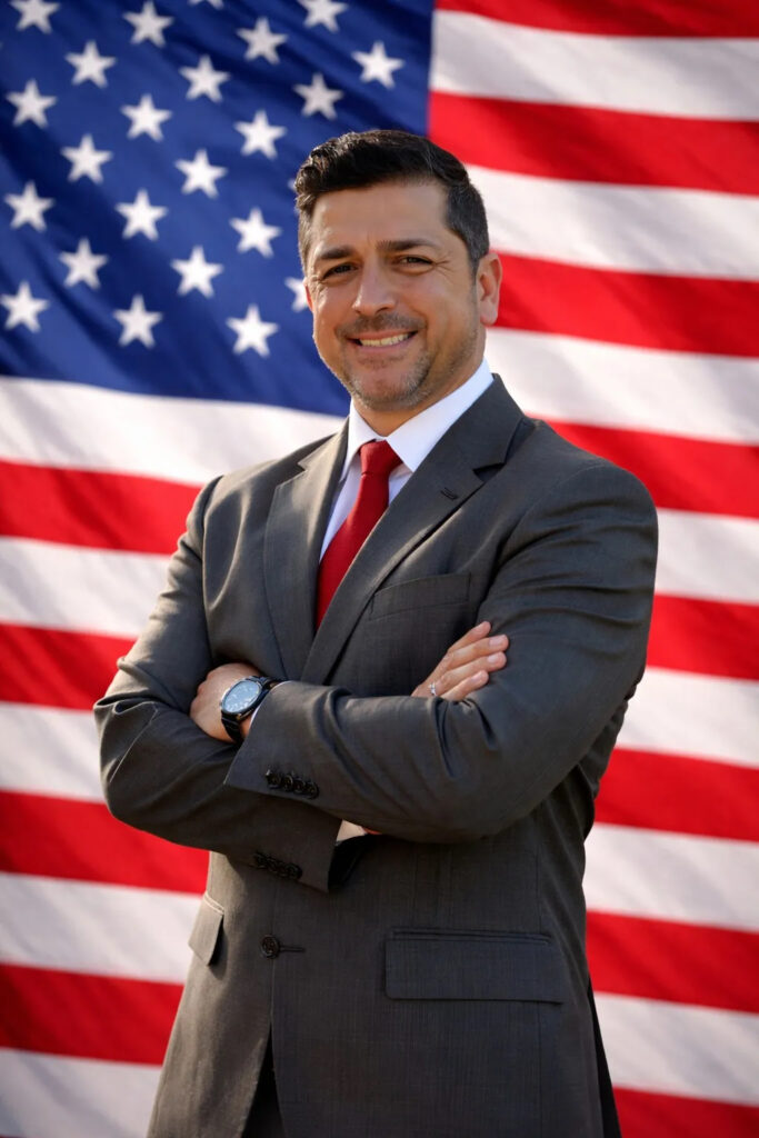 Eddie Pope smiling with arms folded in front of a U.S. flag backdrop.