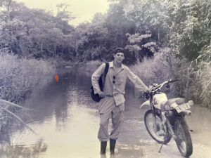 Black and white photo of a young adult Kelly Kirschner with a motorbike near a creek.