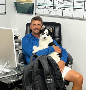 James Peel sits at a desk holding a dog and smiling.