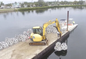 Workers lift reef balls using a crane near the water.