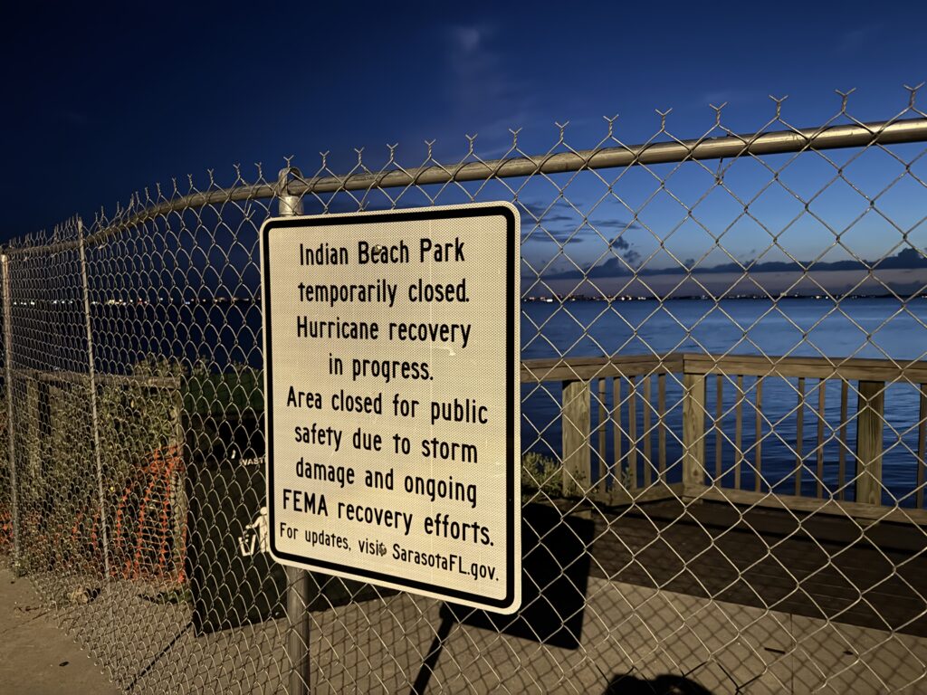 A sign on a chain link fence by the bay reads, "Indian Beach Park temporarily closed. Hurricane recovery in progress. Area closed for public safety due to storm damage and ongoing FEMA recovery efforts. For updates, visit SarasotaFL.gov.