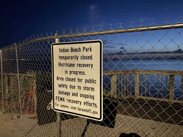 A sign on a chain link fence by the bay reads, "Indian Beach Park temporarily closed. Hurricane recovery in progress. Area closed for public safety due to storm damage and ongoing FEMA recovery efforts. For updates, visit SarasotaFL.gov.