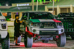 A silver truck with a Mexican flag on the hood. Text on the windshield reads "TACO LIFE".