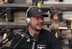 Kyle Rittenhouse speaking in a studio in front of a wall lined with guns.