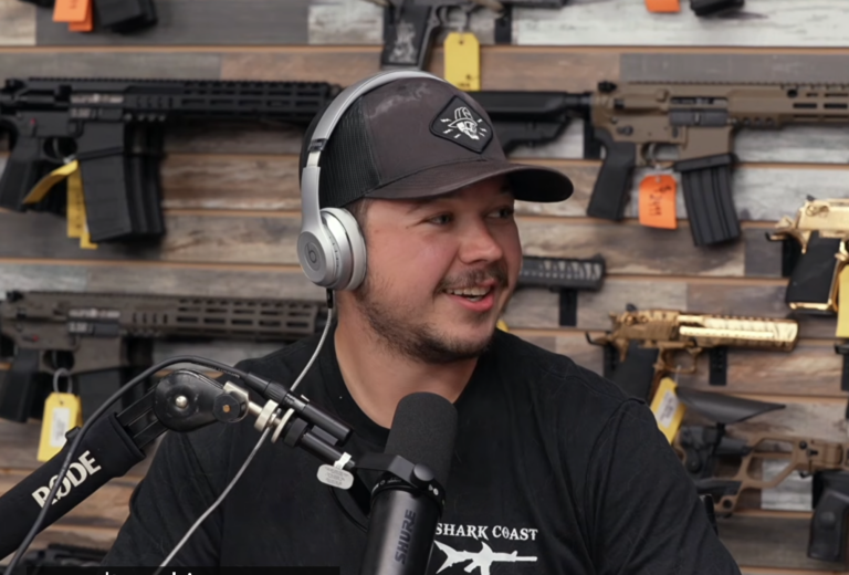Kyle Rittenhouse speaking in a studio in front of a wall lined with guns.