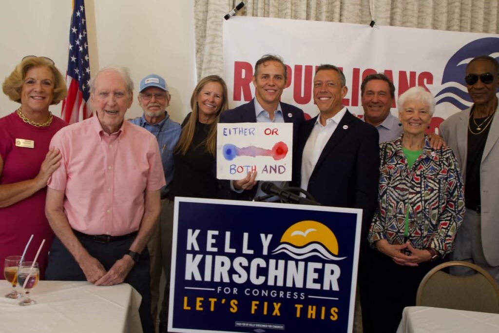 Several smiling people surrund Kelly Kirschner, who holds up a sign with a line fading from blue to purple to red which reads "Either or" on the top and "both and" on the bottom.