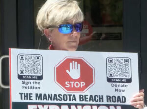 A demonstrator holds a sign that reads "Stop the Manasota Beach Road Expansion" with QR codes labeled "Sign the Petition" and "Donate Now."