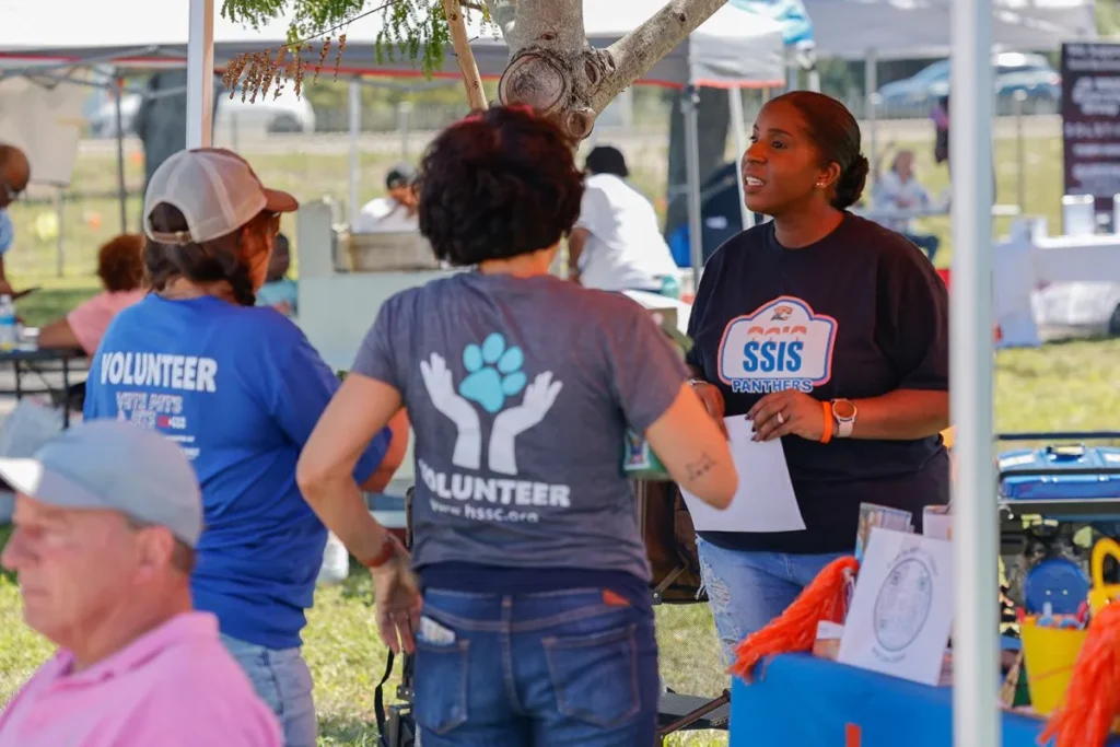 Two people in shirts that say "volunteer" on the back speak to a person with an SSIS Panthers shirt under a tent at an outdoor event.