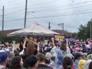 Members of a crowd at an event hold signs in support of Angie Nixon with slogans including "Tax the billionaires" and "Medicare for all".
