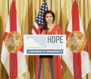 Casey DeSantis speaking behind a Hope Florida podium in front of two Florida flags and a U.S. flag.